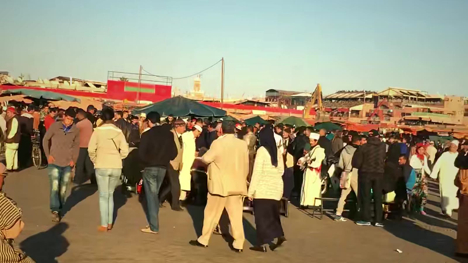 A crowded square in Marrakech in late afternoon light, market umbrellas, and people crossing the open ground.